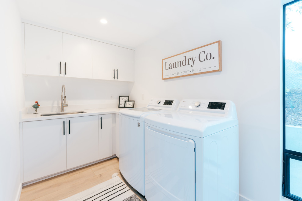 Design ideas for a mid-sized beach style l-shaped dedicated laundry room in Orange County with an undermount sink, flat-panel cabinets, white cabinets, quartz benchtops, white splashback, engineered quartz splashback, white walls, medium hardwood floors, a side-by-side washer and dryer and white benchtop.