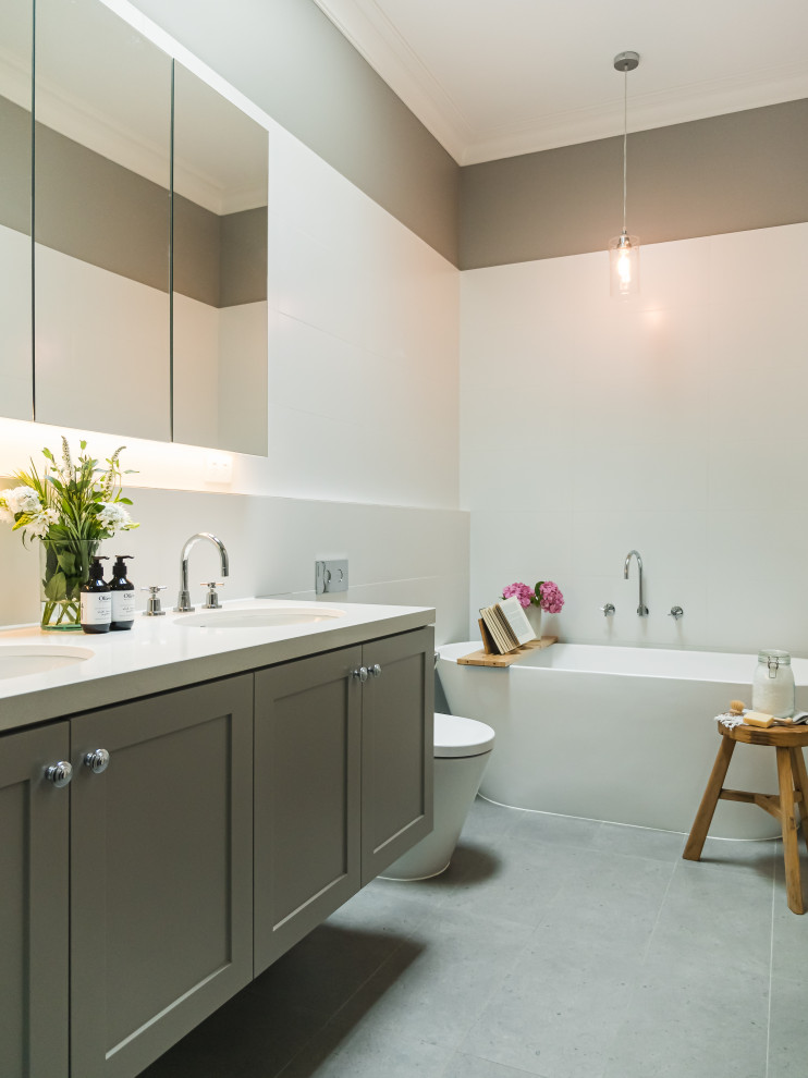 На фото: ванная комната в стиле неоклассика (современная классика) с фасадами в стиле шейкер, серыми фасадами, отдельно стоящей ванной, белой плиткой, серыми стенами, серым полом, белой столешницей, тумбой под две раковины и подвесной тумбой