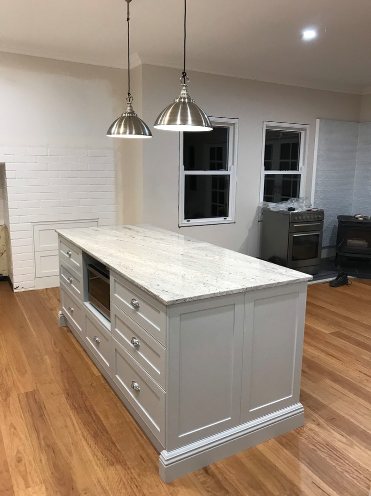 This is an example of a large country eat-in kitchen with a farmhouse sink, shaker cabinets, white cabinets, granite benchtops, white splashback, ceramic splashback, medium hardwood floors, with island and grey benchtop.