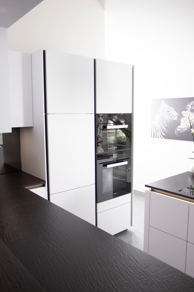 Photo of a small modern u-shaped open plan kitchen in Munich with a single-bowl sink, flat-panel cabinets, white cabinets, black splashback, mirror splashback, black appliances, ceramic floors, with island, grey floor and black benchtop.