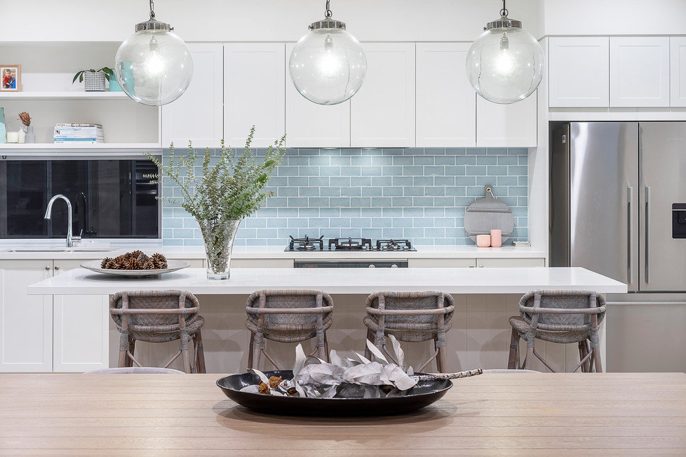 Photo of a mid-sized beach style galley open plan kitchen in Perth with a drop-in sink, shaker cabinets, white cabinets, marble benchtops, blue splashback, subway tile splashback, stainless steel appliances, light hardwood floors, with island, brown floor and white benchtop.