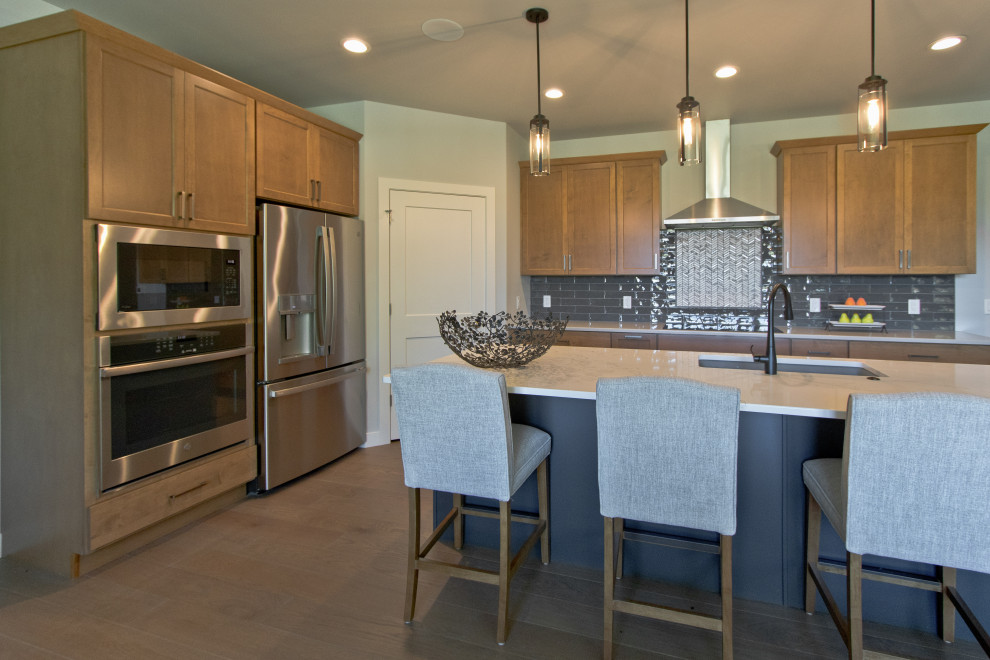 Example of a transitional l-shaped light wood floor and brown floor open concept kitchen design with an undermount sink, shaker cabinets, medium tone wood cabinets, gray backsplash, subway tile backsplash, stainless steel appliances, an island and white countertops