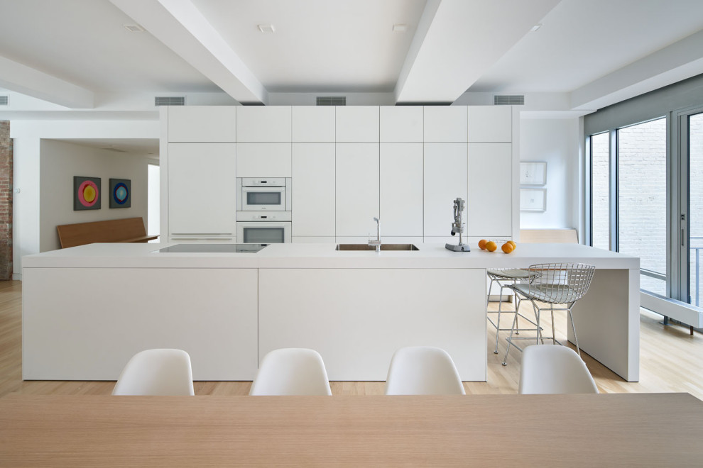 This is an example of a large modern galley eat-in kitchen in New York with a double-bowl sink, flat-panel cabinets, white cabinets, quartzite benchtops, panelled appliances, light hardwood floors, with island, beige floor, white benchtop and exposed beam.