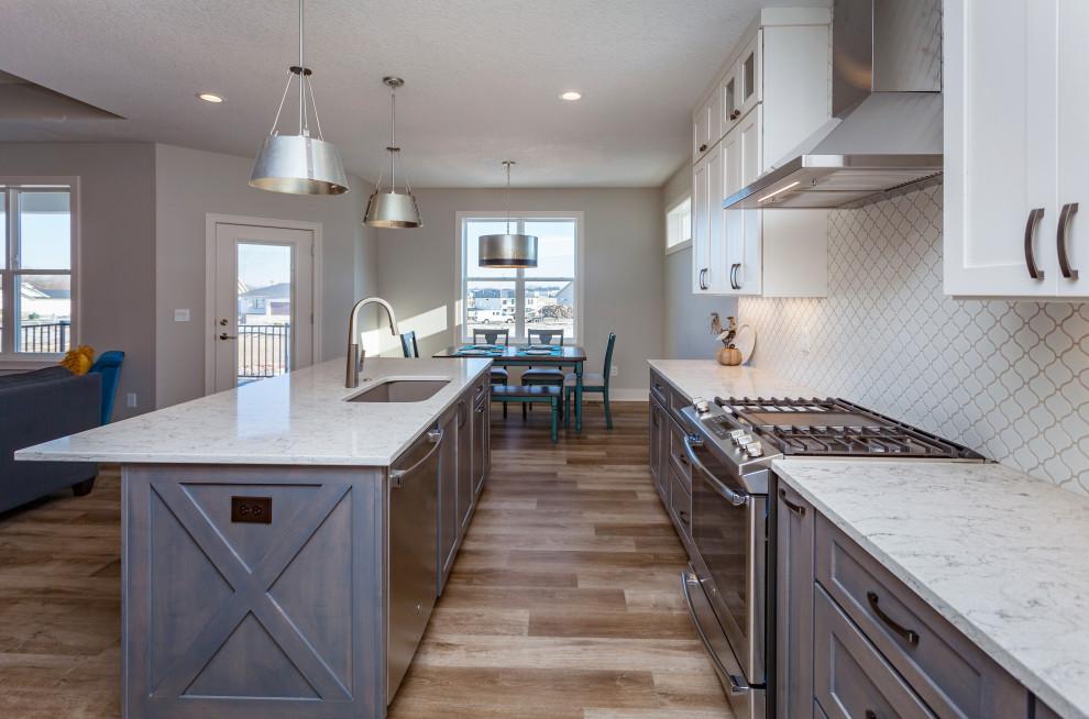Example of a mid-sized transitional l-shaped light wood floor and brown floor open concept kitchen design with an undermount sink, shaker cabinets, white cabinets, quartz countertops, white backsplash, ceramic backsplash, stainless steel appliances, an island and white countertops