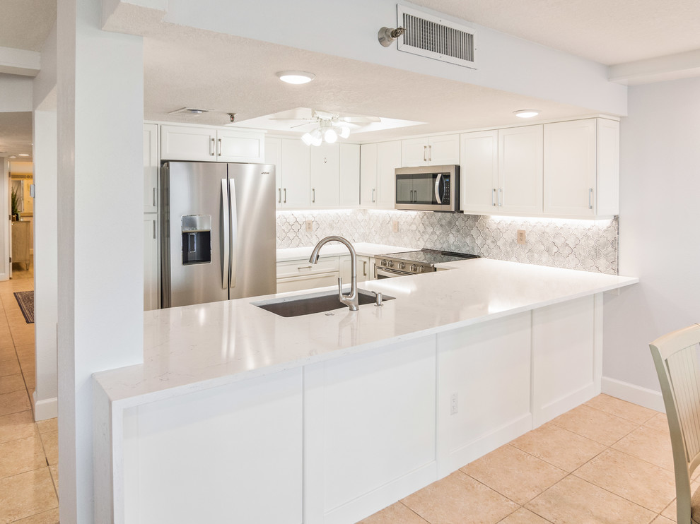 This is an example of a mid-sized beach style u-shaped eat-in kitchen in Tampa with an undermount sink, shaker cabinets, white cabinets, quartzite benchtops, grey splashback, ceramic splashback, stainless steel appliances, porcelain floors, a peninsula, beige floor and white benchtop.