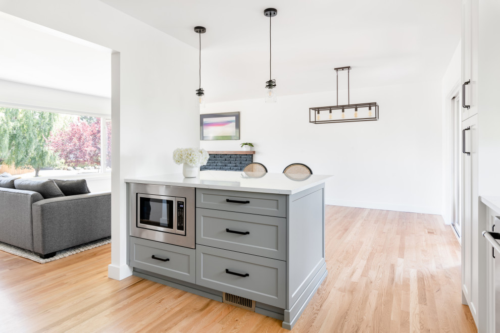 Small classic l-shaped kitchen/diner in Vancouver with a belfast sink, shaker cabinets, white cabinets, engineered stone countertops, white splashback, ceramic splashback, stainless steel appliances, light hardwood flooring, an island, yellow floors and white worktops.
