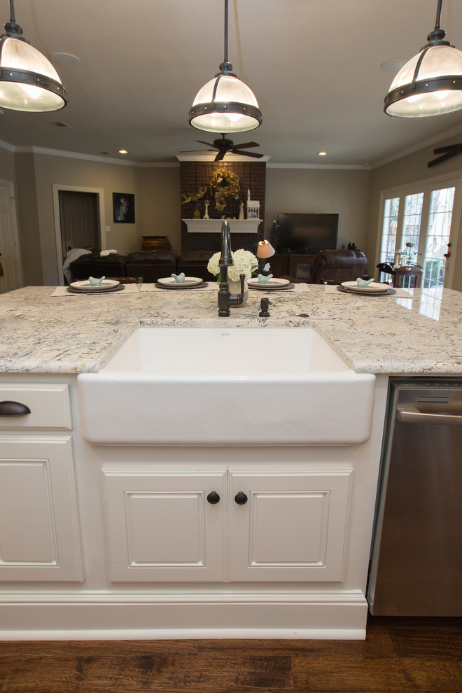 This is an example of a large country single-wall open plan kitchen in Dallas with granite benchtops, with island, a farmhouse sink, raised-panel cabinets, white cabinets, white splashback, subway tile splashback, stainless steel appliances, dark hardwood floors, brown floor and white benchtop.