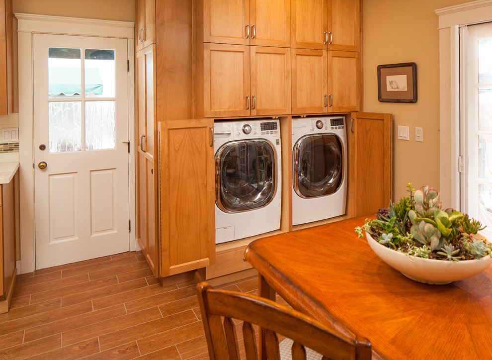 Photo of a large traditional single-wall utility room in San Diego with shaker cabinets, medium wood cabinets, quartz benchtops, beige walls, porcelain floors, a side-by-side washer and dryer and brown floor.