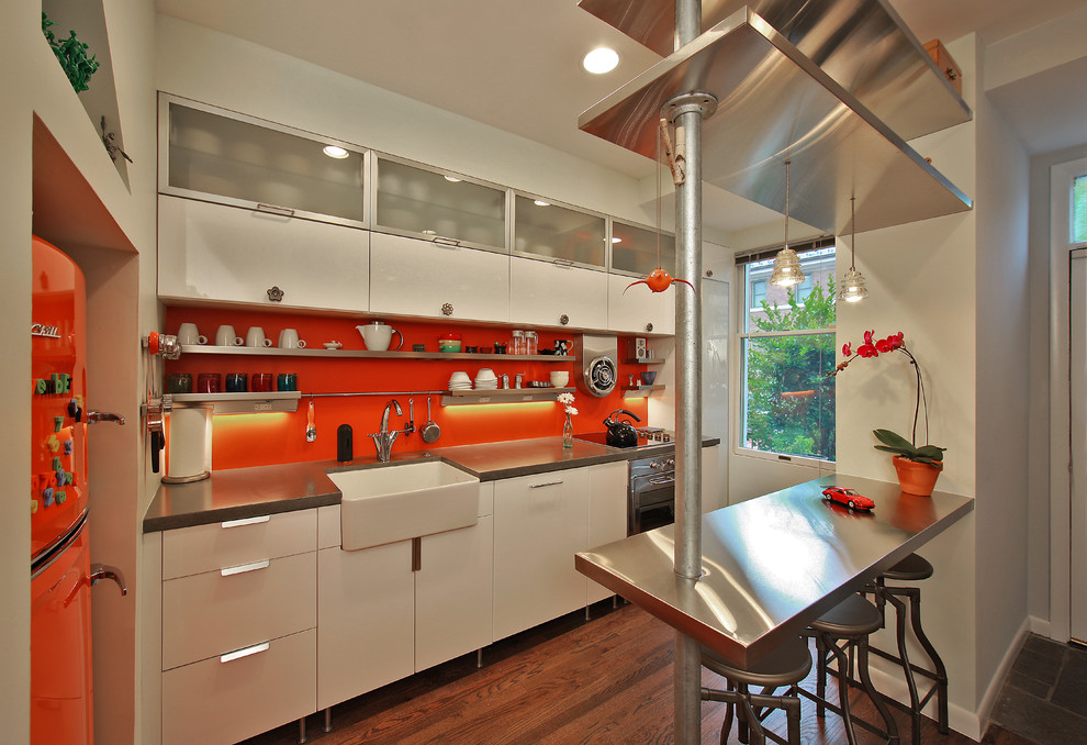 This is an example of a small eclectic galley separate kitchen in DC Metro with a farmhouse sink, flat-panel cabinets, white cabinets, concrete benchtops, orange splashback, stainless steel appliances, medium hardwood floors and brown floor.