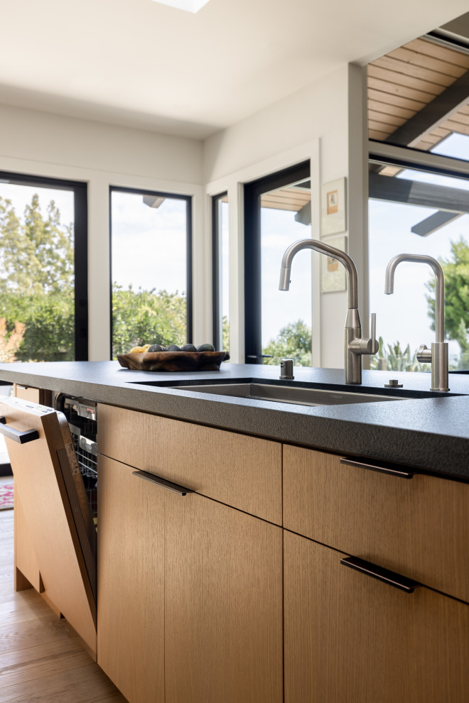 Photo of a large modern l-shaped eat-in kitchen in Santa Barbara with a drop-in sink, flat-panel cabinets, black cabinets, soapstone benchtops, white splashback, marble splashback, stainless steel appliances, light hardwood floors, with island, brown floor and black benchtop.