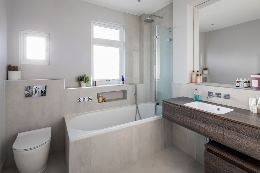 Modern kids bathroom in London with flat-panel cabinets, dark wood cabinets, a drop-in tub, a shower/bathtub combo, a one-piece toilet, white tile, stone slab, white walls, ceramic floors, an integrated sink, wood benchtops, white floor and a hinged shower door.