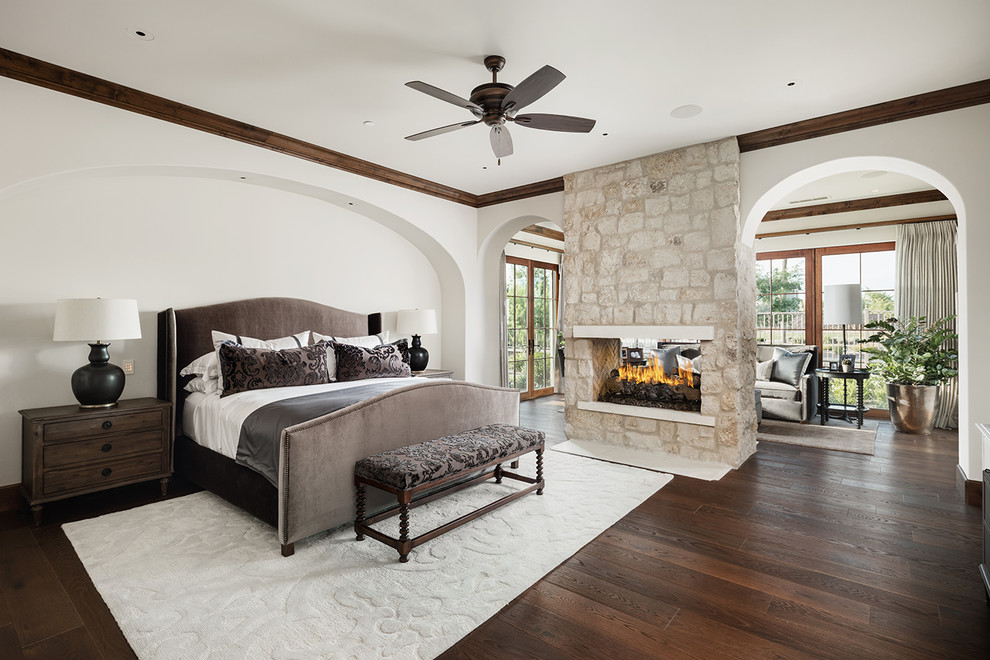 Photo of a mid-sized mediterranean master bedroom in Phoenix with white walls, dark hardwood floors, a two-sided fireplace, a stone fireplace surround and brown floor.
