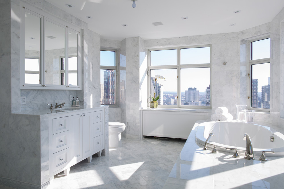 Design ideas for a contemporary bathroom in New York with an undermount sink, recessed-panel cabinets, white cabinets, a drop-in tub, a two-piece toilet, gray tile, stone tile and marble floors.