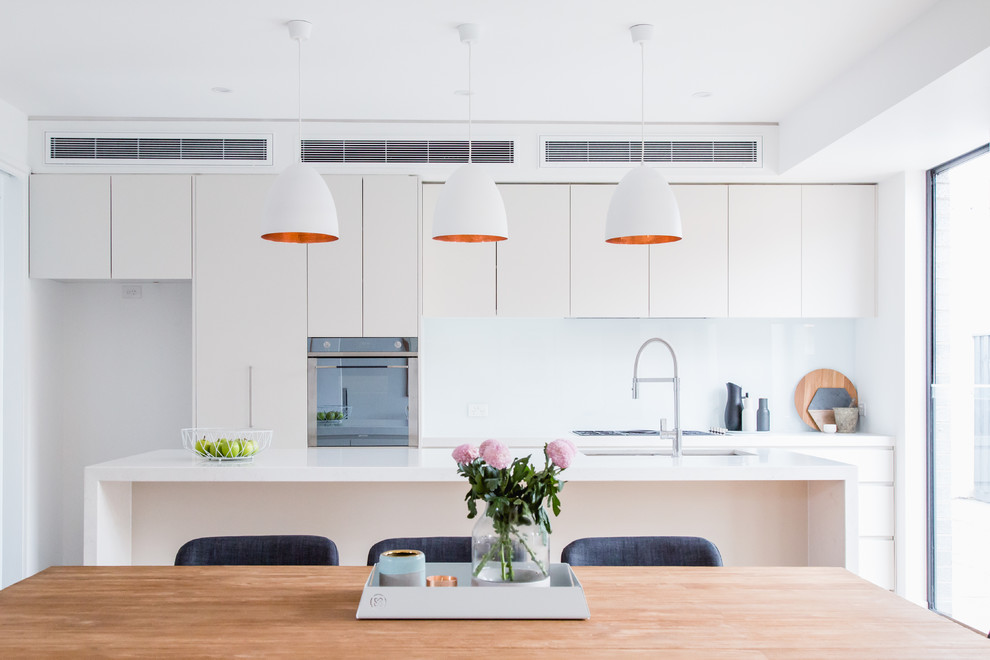 Inspiration for a mid-sized contemporary open plan kitchen in Melbourne with a drop-in sink, flat-panel cabinets, white cabinets, wood benchtops, white splashback, ceramic splashback, white appliances, light hardwood floors, with island and white benchtop.