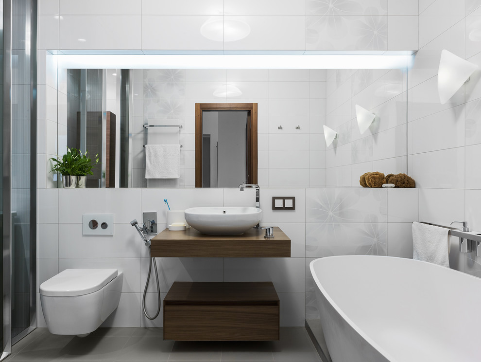 This is an example of a contemporary master bathroom in Moscow with flat-panel cabinets, dark wood cabinets, a freestanding tub, a wall-mount toilet, white tile, a vessel sink and grey floor.