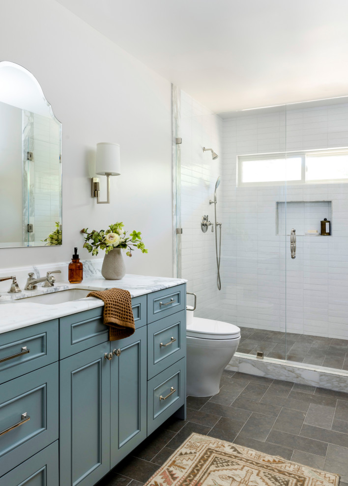 Пример оригинального дизайна: огромная ванная комната в стиле кантри с фасадами с выступающей филенкой, синими фасадами, душем в нише, унитазом-моноблоком, белой плиткой, белыми стенами, полом из известняка, душевой кабиной, накладной раковиной, мраморной столешницей, серым полом, душем с распашными дверями, белой столешницей, сиденьем для душа, тумбой под одну раковину и встроенной тумбой