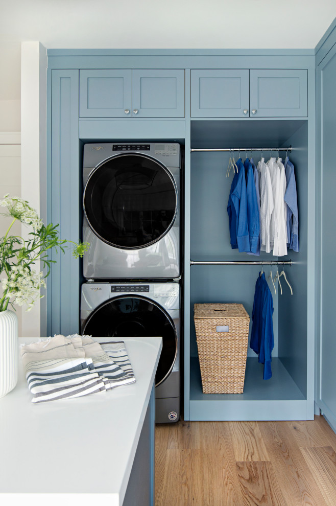 Inspiration for a mid-sized transitional laundry room in Toronto with shaker cabinets, blue cabinets, solid surface benchtops, white walls, light hardwood floors, a stacked washer and dryer and white benchtop.