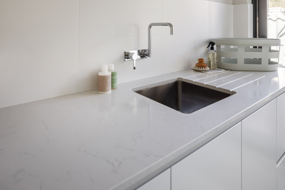 Inspiration for a mid-sized contemporary single-wall utility room in Perth with an undermount sink, flat-panel cabinets, quartz benchtops, white splashback, porcelain splashback, porcelain floors, a stacked washer and dryer, grey floor and white benchtop.