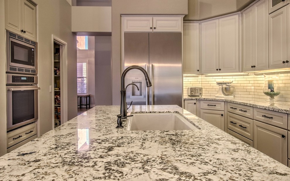 This is an example of a mid-sized eclectic l-shaped eat-in kitchen in Phoenix with an undermount sink, raised-panel cabinets, white cabinets, granite benchtops, white splashback, ceramic splashback, stainless steel appliances, porcelain floors, with island, white floor and white benchtop.