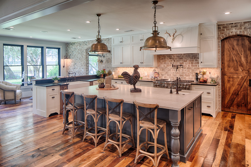 This is an example of a country l-shaped kitchen in Atlanta with a farmhouse sink, shaker cabinets, white cabinets, brick splashback, stainless steel appliances, medium hardwood floors, with island, brown floor and black benchtop.