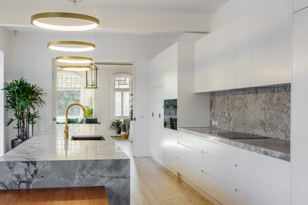 Large contemporary galley eat-in kitchen in Melbourne with an undermount sink, white cabinets, marble benchtops, grey splashback, marble splashback, black appliances, light hardwood floors, with island and grey benchtop.