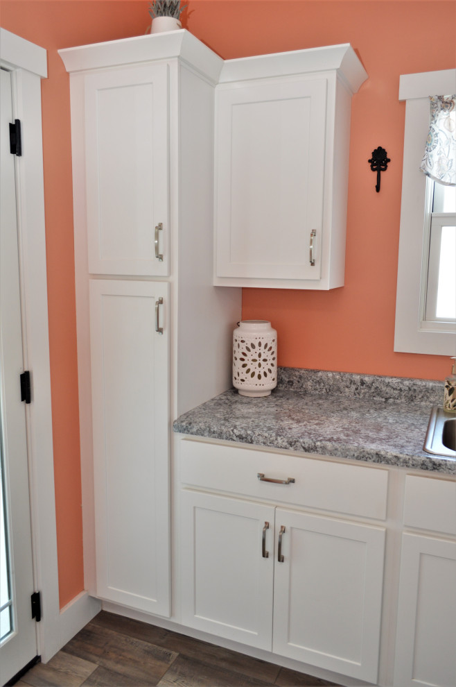 This is an example of a mid-sized country l-shaped dedicated laundry room in Other with a drop-in sink, shaker cabinets, white cabinets, laminate benchtops, pink walls, laminate floors, a side-by-side washer and dryer, brown floor and grey benchtop.