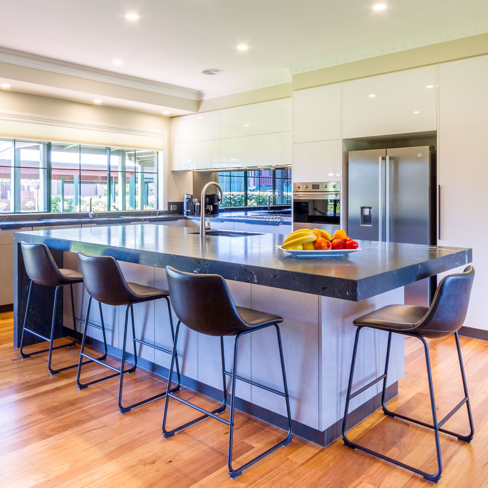 This is an example of a large modern l-shaped open plan kitchen in Melbourne with a double-bowl sink, flat-panel cabinets, grey cabinets, quartzite benchtops, metallic splashback, mirror splashback, stainless steel appliances, medium hardwood floors, with island, brown floor and black benchtop.