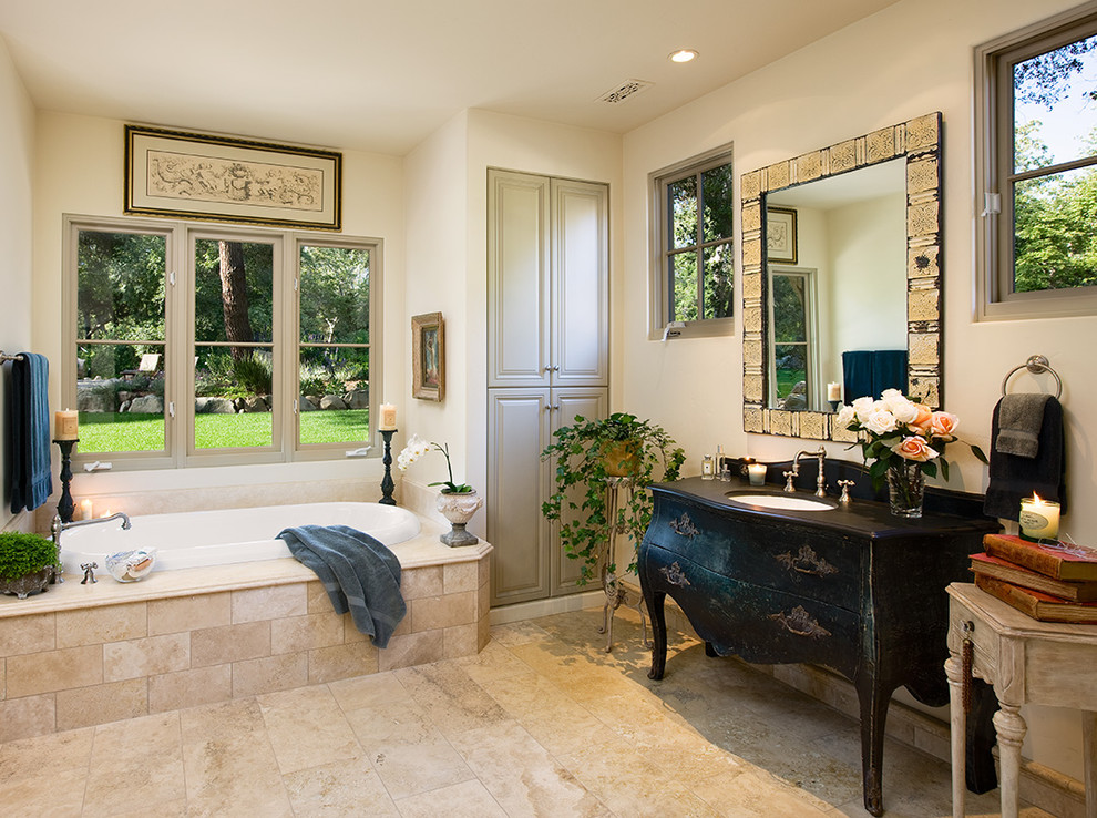 Mediterranean bathroom in Santa Barbara with black cabinets, a drop-in tub, beige tile, travertine and flat-panel cabinets.