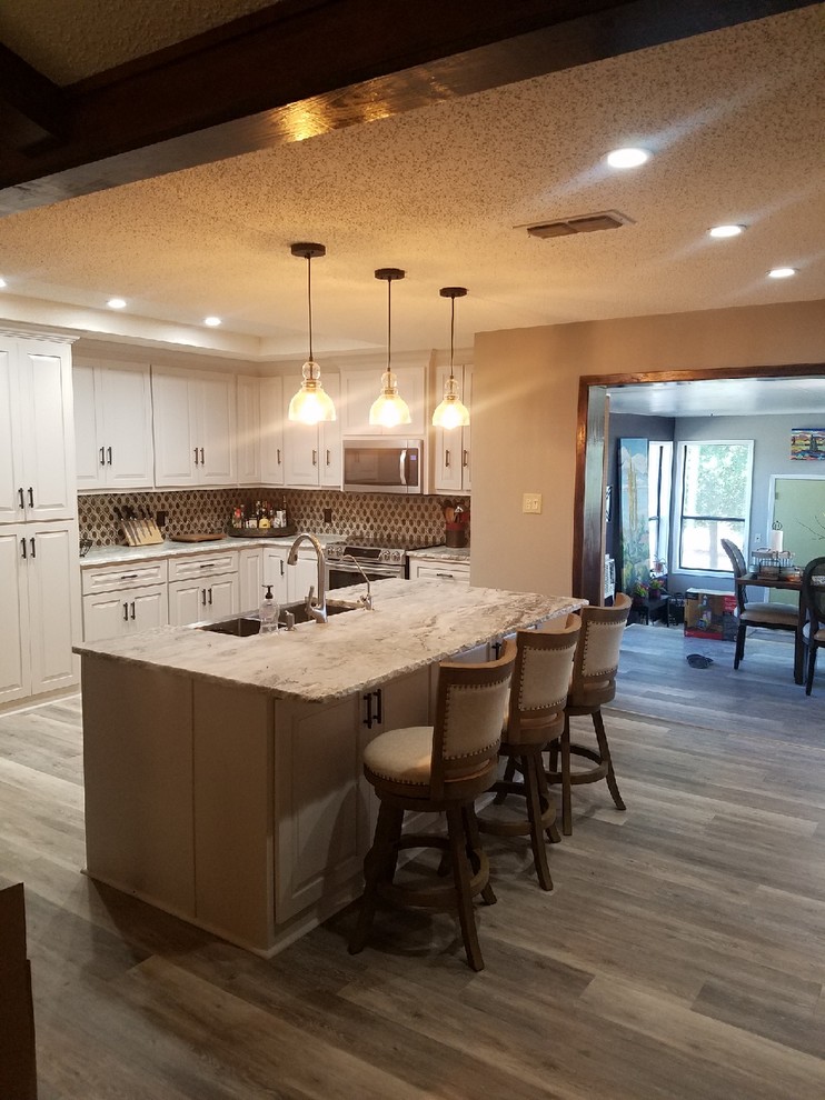 Large transitional l-shaped vinyl floor and brown floor kitchen photo with a farmhouse sink, multicolored backsplash, stainless steel appliances, raised-panel cabinets, white cabinets, granite countertops and an island