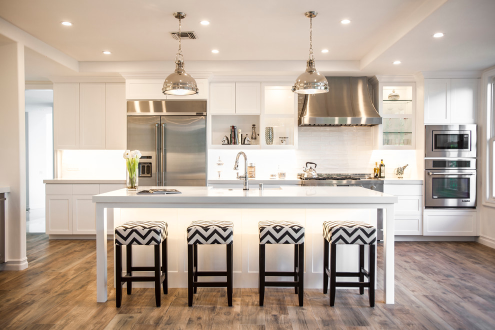 Open concept kitchen - large transitional l-shaped medium tone wood floor open concept kitchen idea with an island, a farmhouse sink, recessed-panel cabinets, white cabinets, quartz countertops, white backsplash, subway tile backsplash and stainless steel appliances