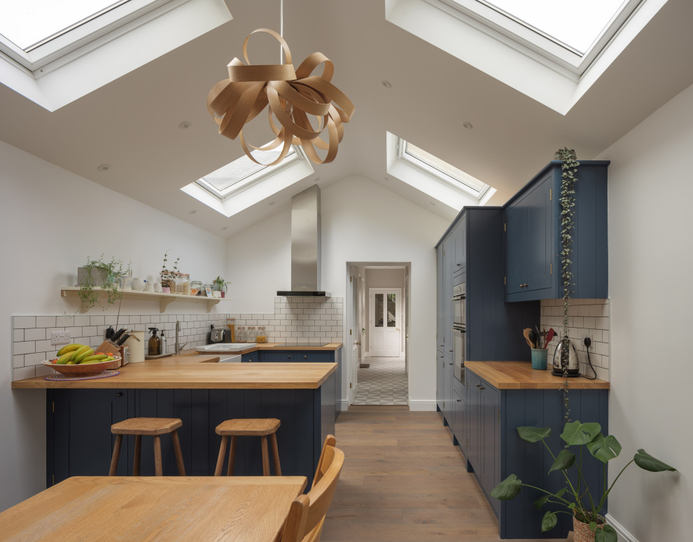Inspiration for a transitional u-shaped kitchen in Cambridgeshire with a farmhouse sink, shaker cabinets, blue cabinets, wood benchtops, white splashback, subway tile splashback, medium hardwood floors, a peninsula, brown floor, beige benchtop and vaulted.