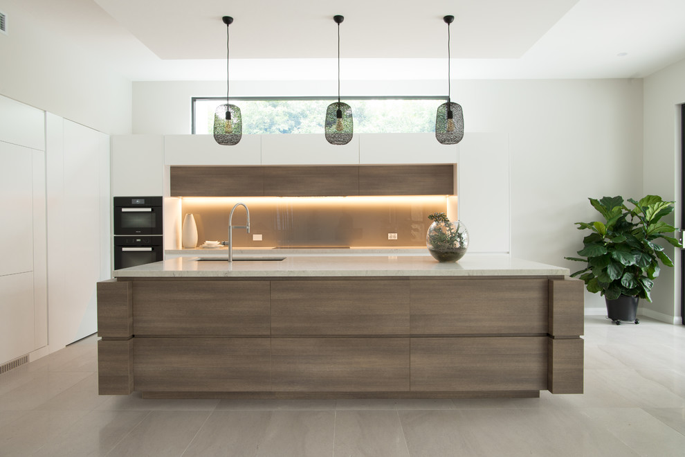 Photo of an expansive contemporary galley open plan kitchen in Sydney with an undermount sink, flat-panel cabinets, white cabinets, quartz benchtops, brown splashback, glass sheet splashback, black appliances, ceramic floors, with island, grey floor and white benchtop.