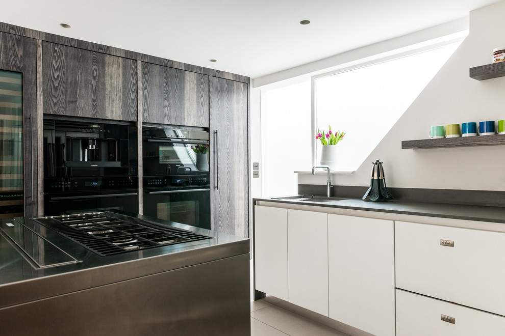 Large contemporary galley open plan kitchen in London with a built-in sink, flat-panel cabinets, white cabinets, composite countertops, white splashback, stone slab splashback, black appliances, slate flooring and multiple islands.