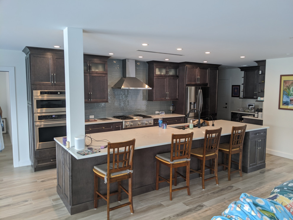 This is an example of a large tropical single-wall open plan kitchen in Miami with a double-bowl sink, recessed-panel cabinets, grey cabinets, quartzite benchtops, blue splashback, glass tile splashback, stainless steel appliances, porcelain floors, with island, brown floor and white benchtop.