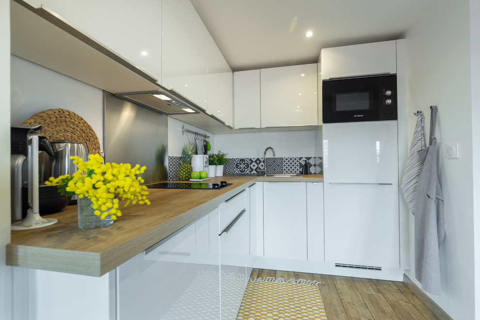 Mid-sized contemporary l-shaped kitchen in Nice with a drop-in sink, white cabinets, wood benchtops, metallic splashback, metal splashback, black appliances, medium hardwood floors and no island.