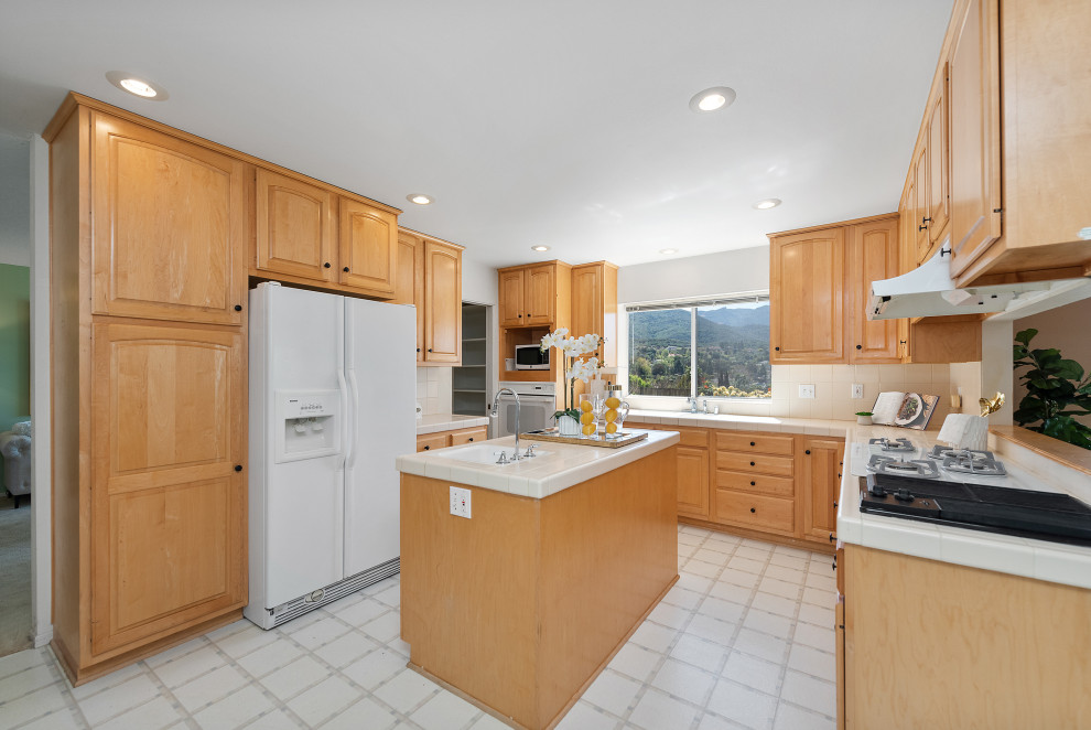 This is an example of a mid-sized country u-shaped eat-in kitchen in Los Angeles with a drop-in sink, raised-panel cabinets, light wood cabinets, tile benchtops, ceramic splashback, white appliances, laminate floors, with island, white floor, white benchtop and wood.