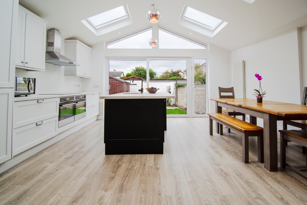 Photo of a mid-sized contemporary galley eat-in kitchen in Dublin with a drop-in sink, louvered cabinets, white cabinets, laminate benchtops, white splashback, black appliances, laminate floors, a peninsula, brown floor and white benchtop.