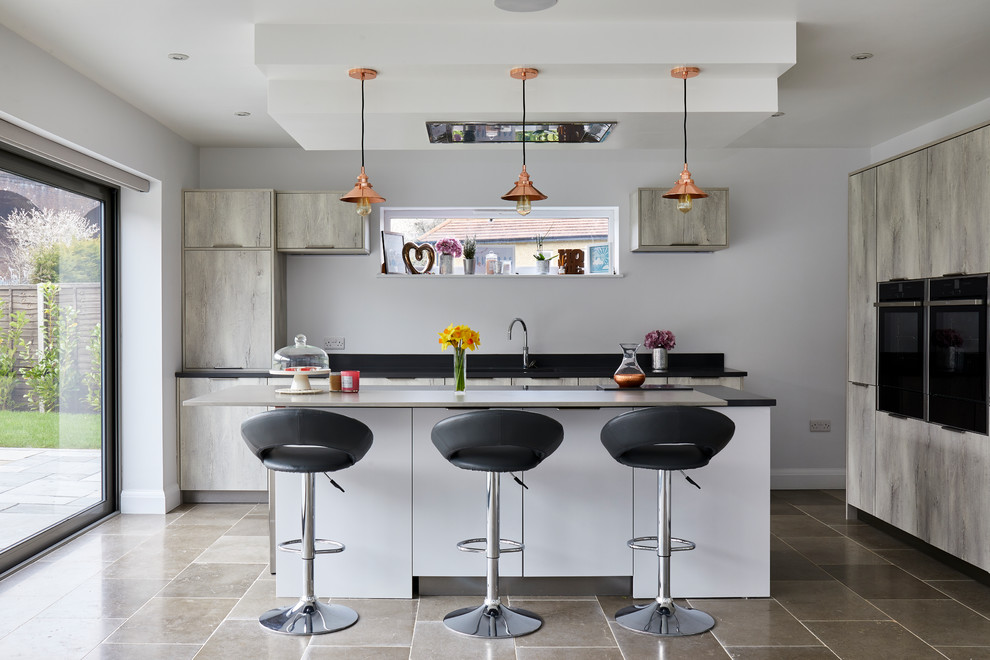 Photo of a contemporary l-shaped kitchen in London with an undermount sink, flat-panel cabinets, light wood cabinets, white splashback, black appliances, with island, grey floor and black benchtop.