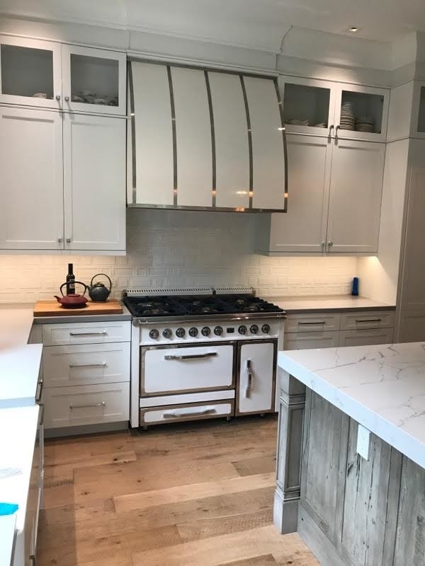 Photo of a mid-sized country l-shaped separate kitchen in Chicago with shaker cabinets, white cabinets, quartz benchtops, white splashback, ceramic splashback, white appliances, medium hardwood floors, with island and brown floor.
