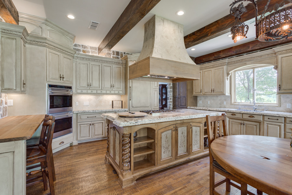 Design ideas for a mid-sized traditional l-shaped eat-in kitchen in Dallas with an undermount sink, raised-panel cabinets, distressed cabinets, granite benchtops, white splashback, travertine splashback, stainless steel appliances, medium hardwood floors, with island and brown floor.