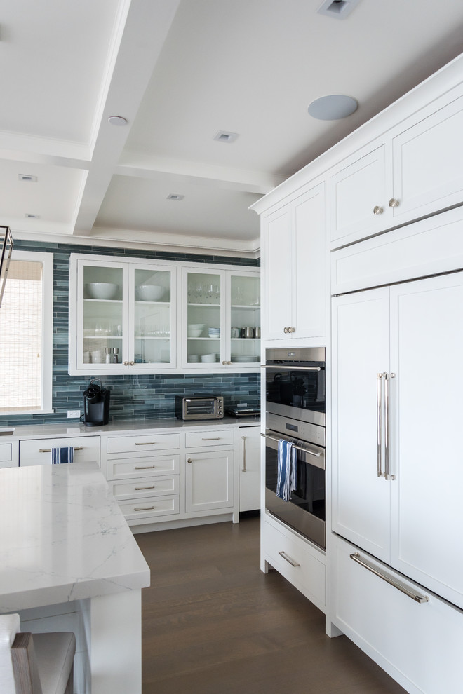 This is an example of a mid-sized beach style single-wall open plan kitchen in New York with an undermount sink, glass-front cabinets, white cabinets, blue splashback, glass tile splashback, stainless steel appliances, medium hardwood floors, with island, brown floor and white benchtop.