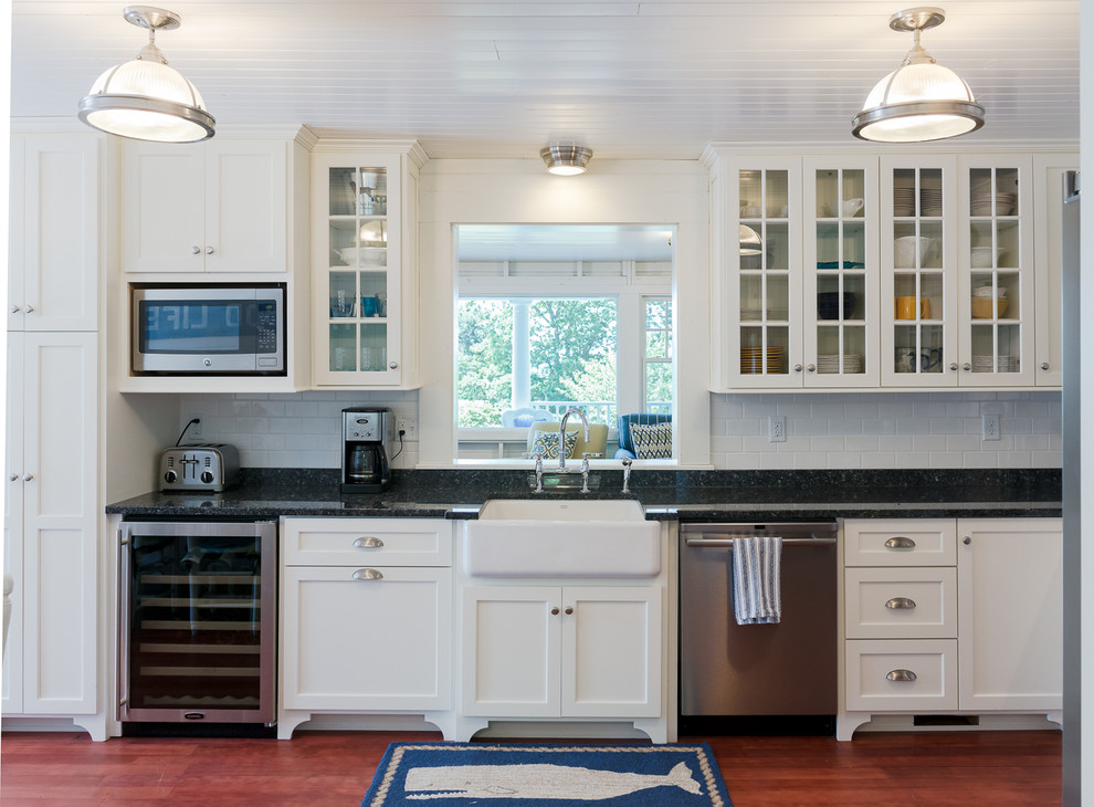 Inspiration for a mid-sized beach style l-shaped eat-in kitchen in Boston with a farmhouse sink, flat-panel cabinets, white cabinets, granite benchtops, white splashback, subway tile splashback, stainless steel appliances, medium hardwood floors and no island.