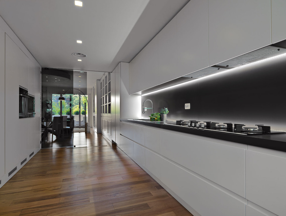 Large contemporary single-wall kitchen in Milan with a drop-in sink, flat-panel cabinets, white cabinets, black splashback, panelled appliances, no island, brown floor and black benchtop.