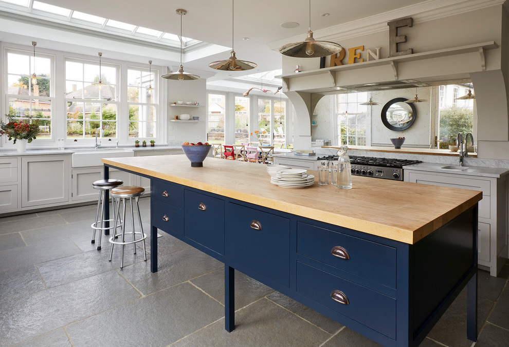 This is an example of a mid-sized country l-shaped eat-in kitchen in Essex with a farmhouse sink, shaker cabinets, mirror splashback, with island, grey floor, blue cabinets, wood benchtops, beige splashback and panelled appliances.