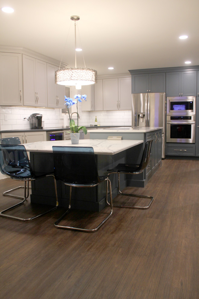 This is an example of a mid-sized transitional l-shaped eat-in kitchen in Other with a single-bowl sink, shaker cabinets, blue cabinets, quartz benchtops, white splashback, ceramic splashback, stainless steel appliances, vinyl floors, multiple islands, brown floor and blue benchtop.