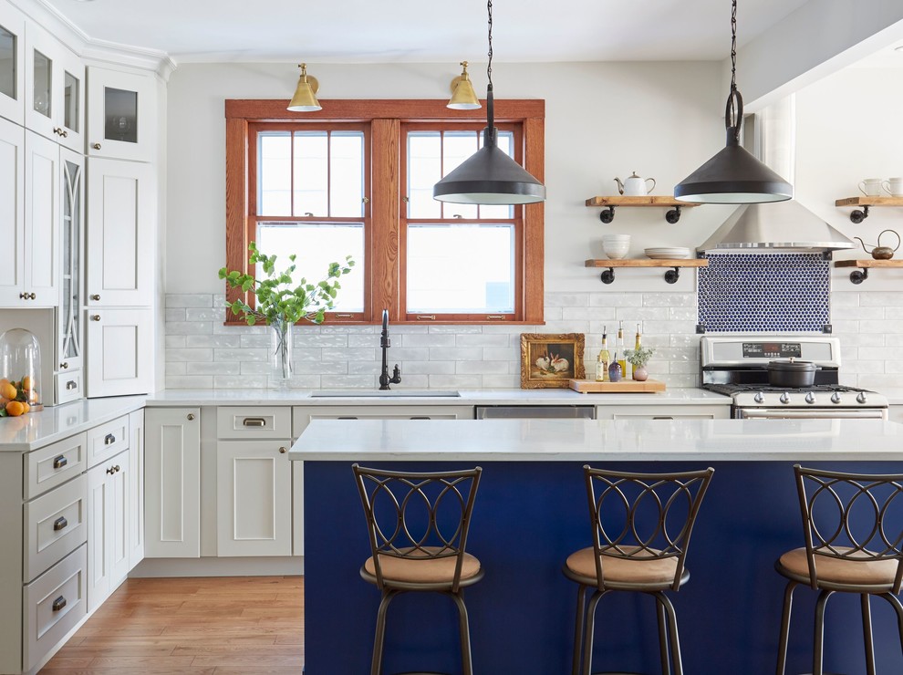 Photo of a traditional l-shaped kitchen in Chicago with an undermount sink, recessed-panel cabinets, white cabinets, white splashback, subway tile splashback, stainless steel appliances, medium hardwood floors, with island, brown floor and white benchtop.