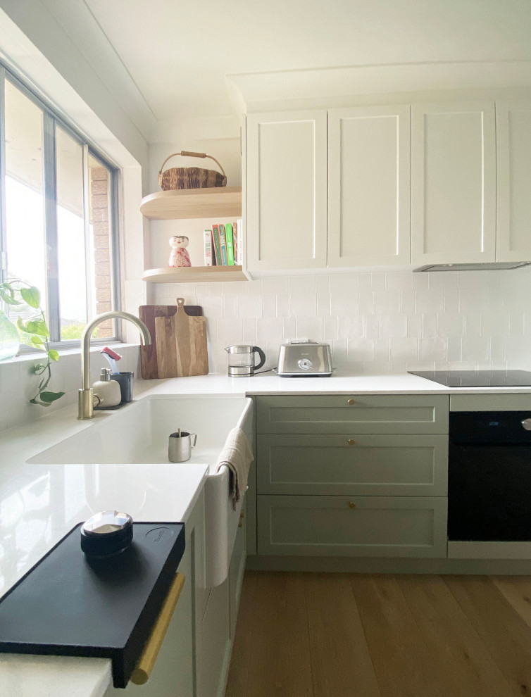 This is an example of a mid-sized modern u-shaped kitchen pantry in Sydney with a farmhouse sink, shaker cabinets, green cabinets, quartz benchtops, white splashback, porcelain splashback, black appliances, light hardwood floors, no island, brown floor and white benchtop.
