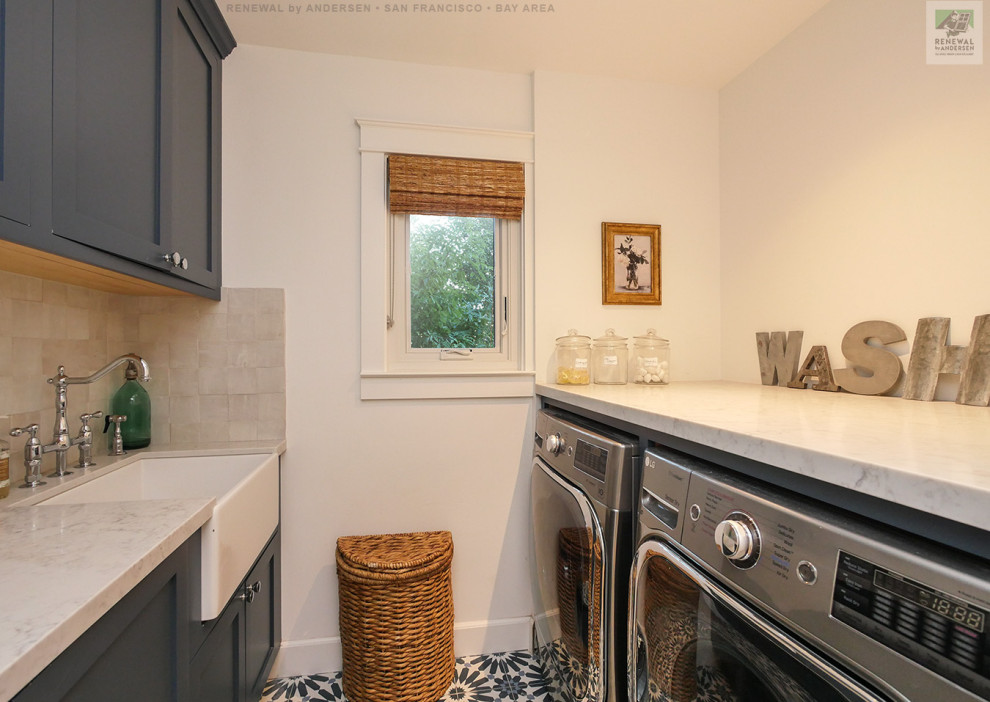 Design ideas for a mid-sized dedicated laundry room in San Francisco with a farmhouse sink, flat-panel cabinets, blue cabinets, granite benchtops, beige splashback, ceramic splashback, white walls, ceramic floors, a side-by-side washer and dryer, blue floor and white benchtop.