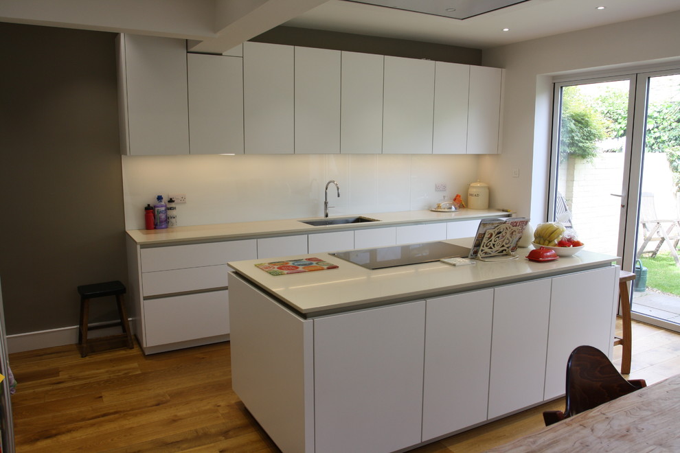 Design ideas for a mid-sized contemporary galley eat-in kitchen in London with a single-bowl sink, flat-panel cabinets, white cabinets, quartzite benchtops, white splashback, glass sheet splashback, black appliances, light hardwood floors and with island.
