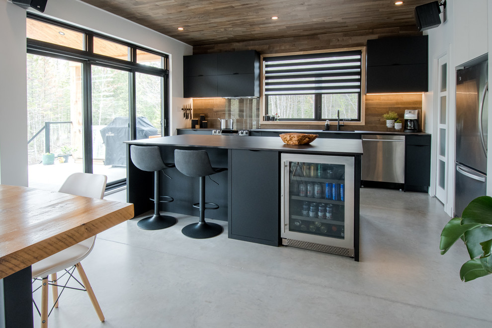 Industrial galley kitchen in Montreal with flat-panel cabinets, black cabinets, brown splashback, stainless steel appliances, concrete floors, with island, grey floor and black benchtop.